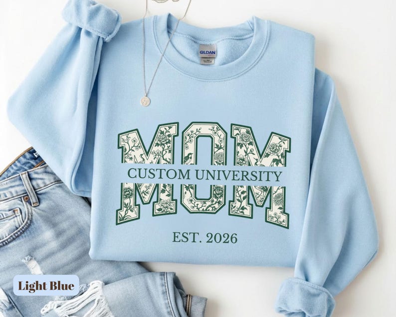May include: A light blue crewneck sweatshirt featuring the word MOM in large block letters with a dark green floral pattern inside. The text CUSTOM UNIVERSITY is printed horizontally across the center, with EST. 2026 printed below in dark green.