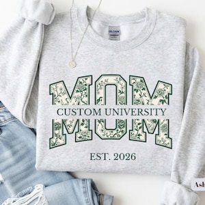 May include: A light gray crewneck sweatshirt featuring the word MOM in large block letters with a dark green floral pattern. The text CUSTOM UNIVERSITY is printed horizontally across the center, with EST. 2026 positioned below in dark green.