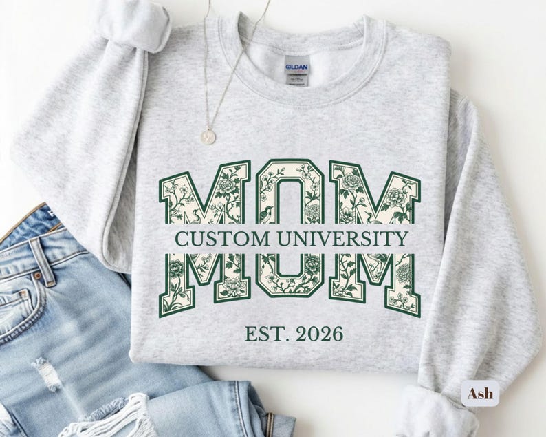May include: A light gray crewneck sweatshirt featuring the word MOM in large block letters with a dark green floral pattern. The text CUSTOM UNIVERSITY is printed horizontally across the center, with EST. 2026 positioned below in dark green.