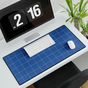 May include: A blue grid pattern desk mat with a white keyboard and mouse. A black computer monitor displays the time as 2:16 PM. The desk is white, and a plant is visible in the background.