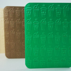 Puede incluir: Tres posavasos de color verde, marrón y crema con un patrón repetido de tazas de café. Las tazas están delineadas en blanco y tienen vapor saliendo de la parte superior.