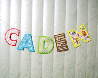 Op de afbeelding: Een kleurrijke banner die de naam "CADEN" spelt met verschillende gepatroneerde letters. De letters zijn gemaakt van papier en aan elkaar geregen met een wit touwtje.