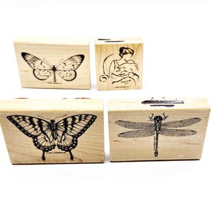 May include: Four wooden stamps with insect and figure designs. The stamps feature a butterfly, a dragonfly, and a woman with a child. The stamps are made of light-colored wood and have black ink designs.