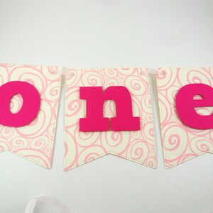 May include: Pink and white banner with the word "one" in large, hot pink letters. The banner is decorated with a swirling pink pattern.