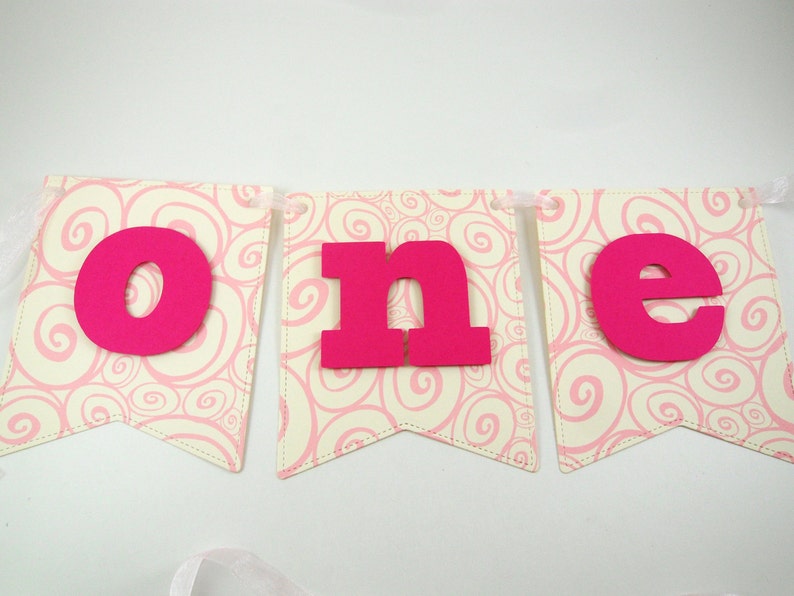 May include: Pink and white banner with the word "one" in large, hot pink letters. The banner is decorated with a swirling pink pattern.