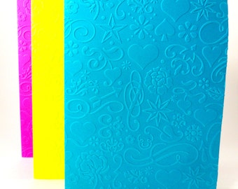 May include: Three notebooks with different colored covers. The front cover is a bright blue with a white embossed pattern of hearts, stars, and swirls. The middle cover is yellow and the back cover is pink.