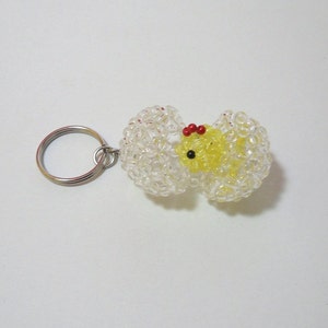 May include: A yellow chick keychain made of beads, with red and black accents. The chick is inside a clear beaded egg, with a silver key ring attached.