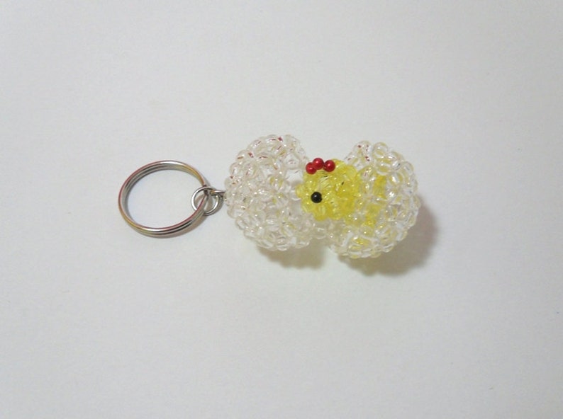 May include: A yellow chick keychain made of beads, with red and black accents. The chick is inside a clear beaded egg, with a silver key ring attached.