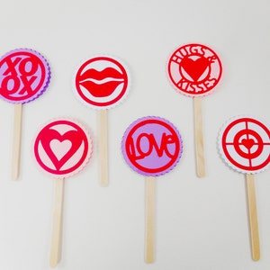May include: Six Valentine's Day themed paper cutouts on sticks. The cutouts are circles with different designs, including hearts, lips, and the words "XOX", "LOVE", and "HUGS & KISSES".