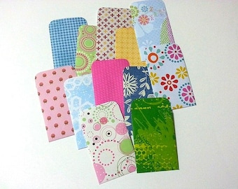 May include: A collection of eleven rectangular paper envelopes featuring various colorful patterns including houndstooth, floral, polka dots, geometric shapes, and solid pink. The envelopes are arranged in an overlapping cluster on a white surface.