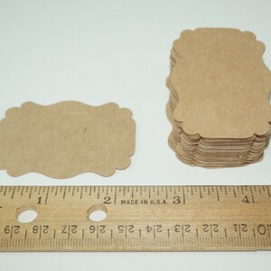May include: A stack of 50 brown kraft paper gift tags with scalloped edges. The tags are approximately 2 inches wide and 3 inches tall. The tags are shown next to a ruler for size reference.