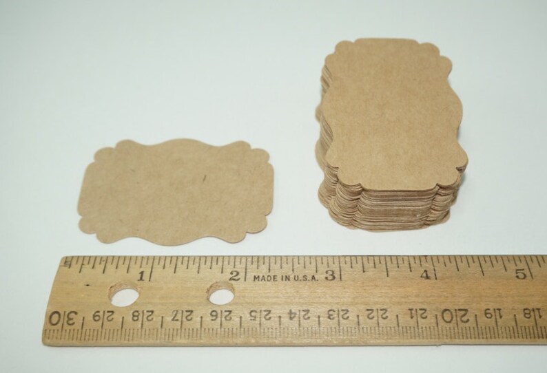 May include: A stack of 50 brown kraft paper gift tags with scalloped edges. The tags are approximately 2 inches wide and 3 inches tall. The tags are shown next to a ruler for size reference.