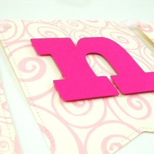 May include: Pink letter "N" banner with a white background featuring a pink swirl pattern. The letter is made of thick cardstock and is attached to the banner with a white ribbon.