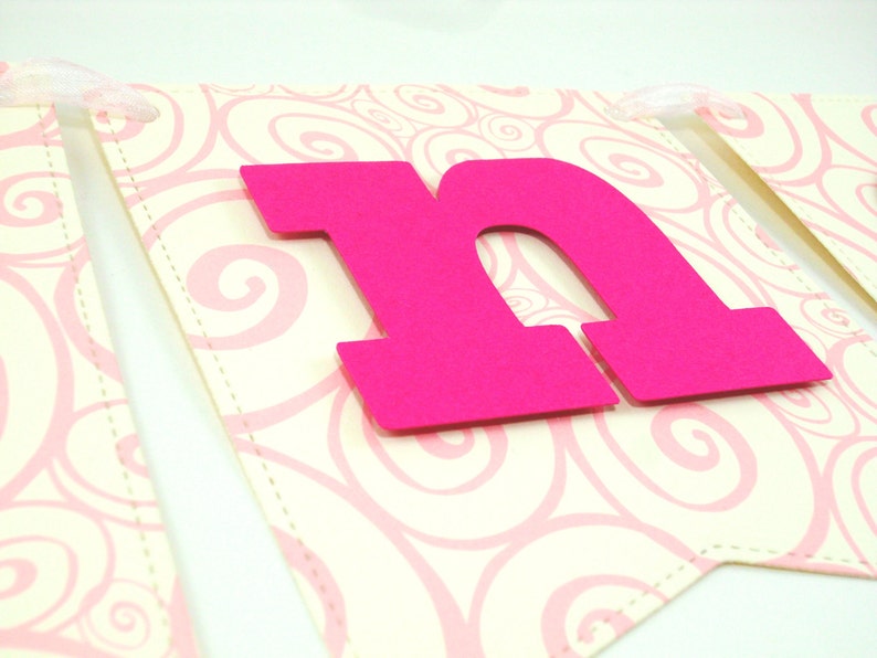 May include: Pink letter "N" banner with a white background featuring a pink swirl pattern. The letter is made of thick cardstock and is attached to the banner with a white ribbon.