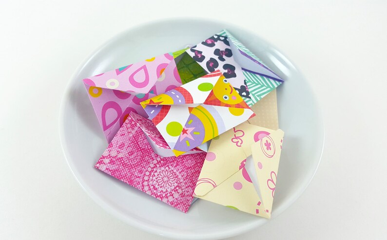 May include: A collection of colorful origami envelopes in a white bowl. The envelopes feature various patterns, including pink paisley, leopard print, and a cartoon face. The bowl is white and sits on a white surface.
