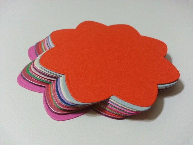Pode incluir: Uma pilha de recortes de papel coloridos em forma de flor. O recorte superior &eacute; laranja, e o resto dos recortes s&atilde;o de v&aacute;rios tons de rosa, roxo, azul e verde.