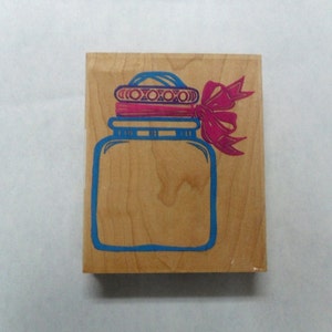 Op de afbeelding: Houten stempel met een blauw en roze ontwerp van een pot met een deksel en een roze strik.