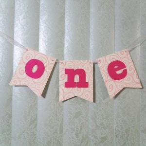 May include: Pink and white banner with the word "one" in pink letters. The banner is decorated with a swirl pattern.