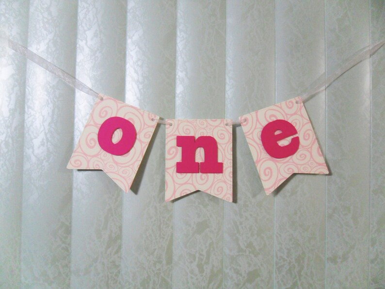 May include: Pink and white banner with the word "one" in pink letters. The banner is decorated with a swirl pattern.