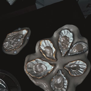 Könnte beinhalten: Dekorative Austern-Servierplatte mit sechs einzelnen Austernschalen-förmigen Fächern. Die Schalen sind silberfarben mit goldenen Verzierungen und kleinen Perlenakzenten. Die Platte ist grau und liegt auf einer schwarzen Oberfläche.
