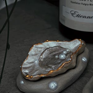 May include: A decorative oyster shell-shaped dish with a gold rim, filled with faux pearls. The dish sits on a gray, oval-shaped base, with additional pearls scattered around. The shell interior has a metallic silver and brown finish.