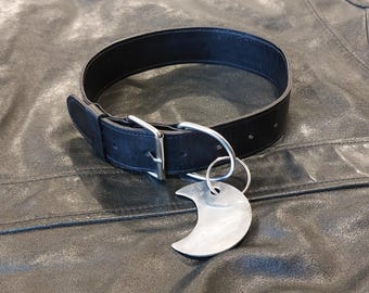 May include: A black leather collar featuring a silver-toned metal buckle and a D-ring. Attached to the D-ring is a circular silver-toned metal pendant shaped like a crescent moon. The collar has a smooth texture and a simple, functional design.