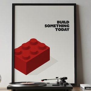 Puede incluir: Una impresión enmarcada con un gran bloque de construcción rojo y el texto "BUILD SOMETHING TODAY" en negro. Un tocadiscos negro se encuentra debajo de la obra de arte en una estantería de madera.