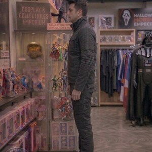 May include: A person stands in a cosplay and collectibles emporium. The store features display cases filled with action figures and collectibles. The person is wearing a dark jacket and pants. The store has a Halloween section with costumes.