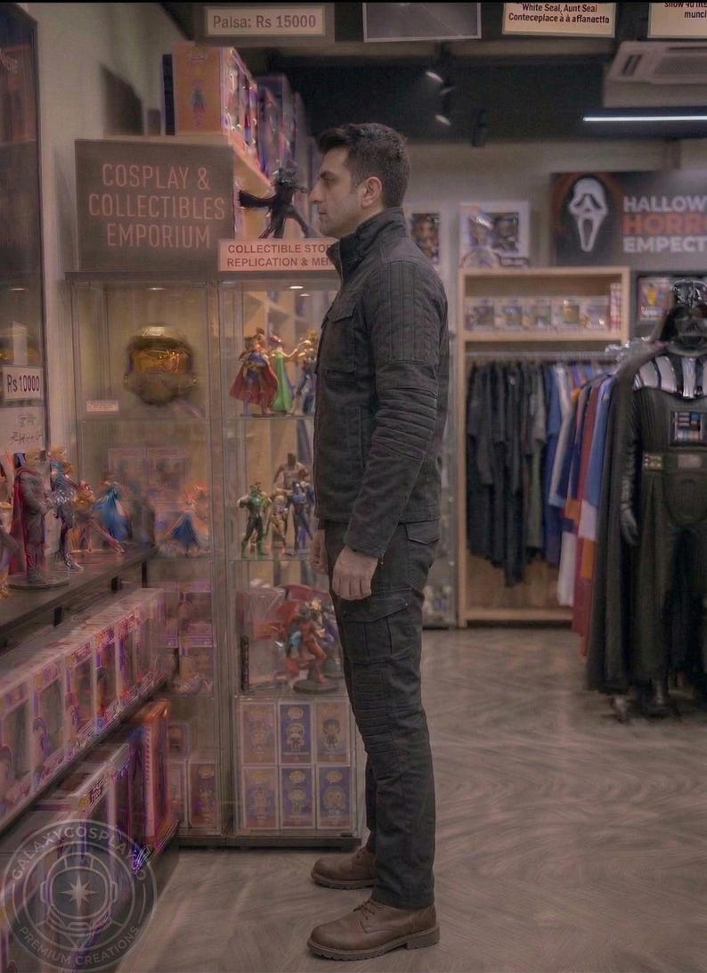 May include: A person stands in a cosplay and collectibles emporium. The store features display cases filled with action figures and collectibles. The person is wearing a dark jacket and pants. The store has a Halloween section with costumes.