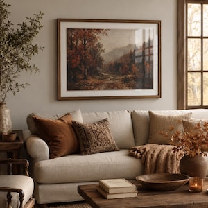 May include: A framed landscape painting depicting an autumn forest path with orange and brown foliage, hanging above a beige sofa with various textured throw pillows, a wooden coffee table with books, a bowl, and a candle in a warm-toned room.