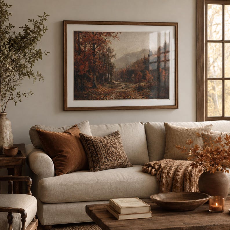 May include: A framed landscape painting depicting an autumn forest path with orange and brown foliage, hanging above a beige sofa with various textured throw pillows, a wooden coffee table with books, a bowl, and a candle in a warm-toned room.