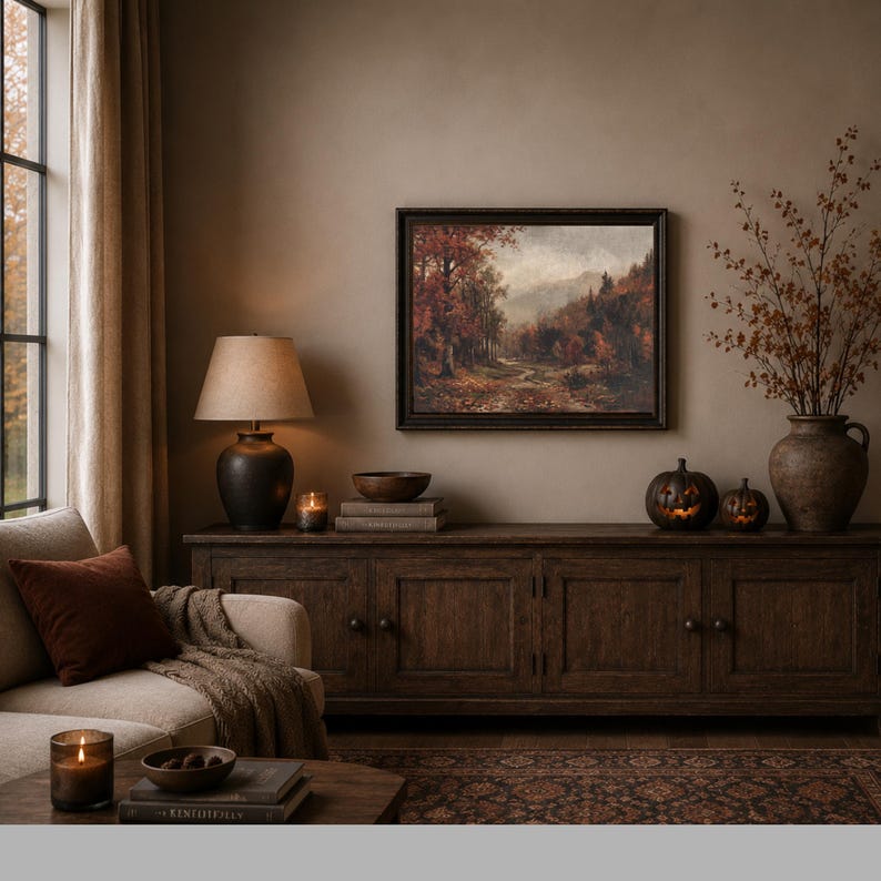 May include: A framed landscape painting depicting an autumn forest path with orange and brown foliage, hanging above a dark wooden sideboard. The scene includes a table lamp, two carved pumpkin decorations, and a ceramic vase with dried branches.