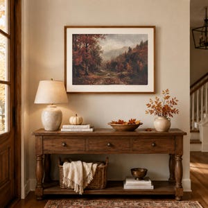 May include: A wooden console table with three drawers and a lower shelf, featuring a table lamp, a small white pumpkin, a bowl of autumn leaves, and a vase of branches. A framed landscape painting of a forest hangs on the wall above the table.