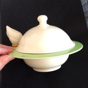 May include: A white ceramic butter dish with a lid and a light green base. The lid has a small knob and a duck wing detail. The dish is designed for butter or spreads. Measurements are in centimetres.