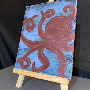 May include: An original painting of a maroon octopus on a light blue background, displayed on a small wooden easel. The octopus has multiple curling tentacles. The background has a textured, brushstroke effect.