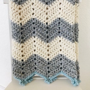May include: A crocheted baby blanket with a chevron pattern in gray, white, and light blue. The blanket has a scalloped edge.