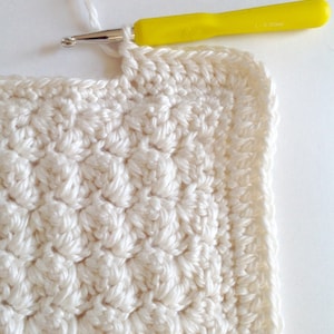 May include: A close-up of a white crocheted square with a yellow crochet hook. The hook is resting on the edge of the square, and the yarn is attached to the hook.