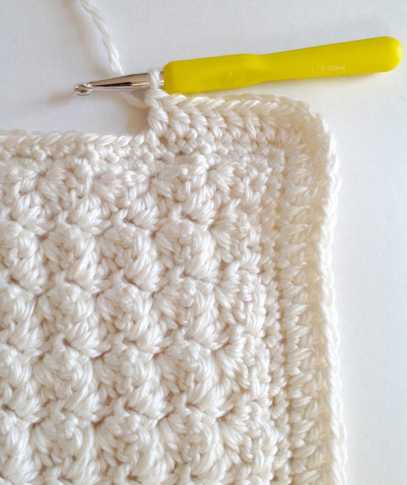 May include: A close-up of a white crocheted square with a yellow crochet hook. The hook is resting on the edge of the square, and the yarn is attached to the hook.