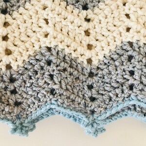 May include: A crocheted baby blanket with a chevron pattern in shades of white, gray, and blue. The blanket has a scalloped edge.