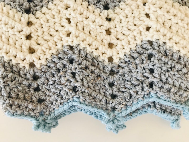 May include: A crocheted baby blanket with a chevron pattern in shades of white, gray, and blue. The blanket has a scalloped edge.