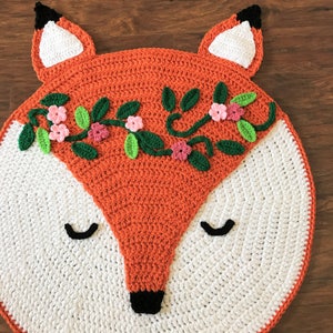 May include: A crocheted fox rug with a white background and orange face. The fox has a floral wreath around its head with pink and green flowers.