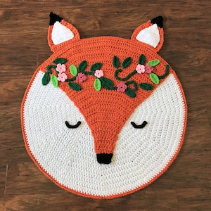 May include: A crocheted fox rug with a white background and orange face. The fox has a floral crown with pink and green accents.