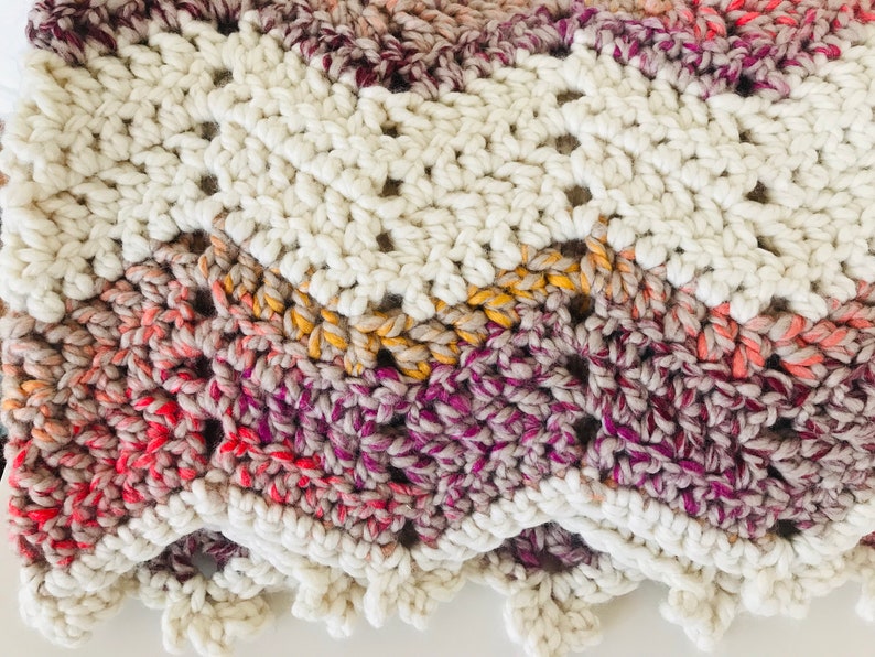 May include: A crocheted blanket with a white border and a multi-colored center. The center is made of a gradient yarn in shades of pink, orange, yellow, and purple.