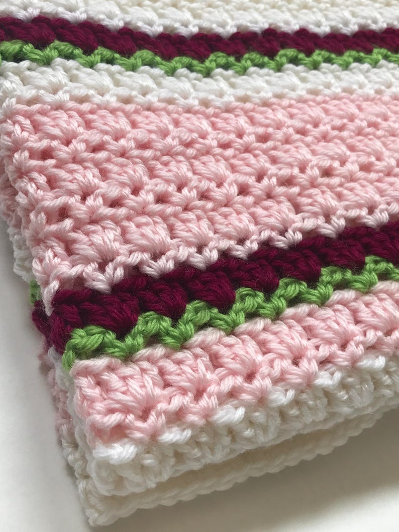 May include: A pink, white, burgundy, and green striped crocheted blanket. The blanket is made with a textured stitch and has a fringed edge.