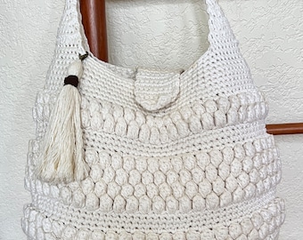 May include: A white crocheted tote bag with a tassel. The bag has a flap closure and a long strap.