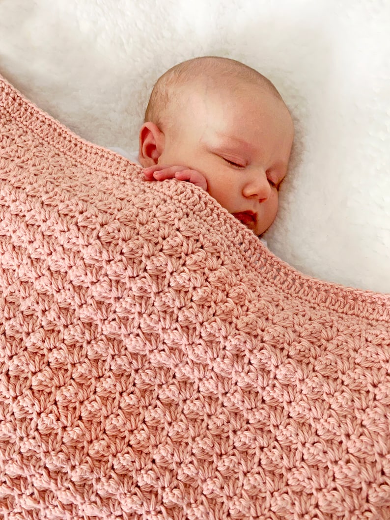 May include: A soft, pink crocheted baby blanket with a delicate stitch pattern. The blanket is draped over a sleeping baby.