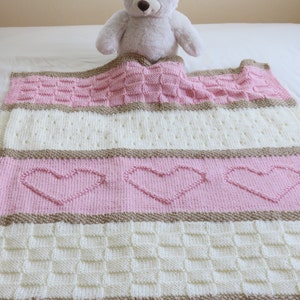 May include: A pink and white knitted baby blanket with a brown stripe and heart pattern. A white teddy bear is sitting on top of the blanket.