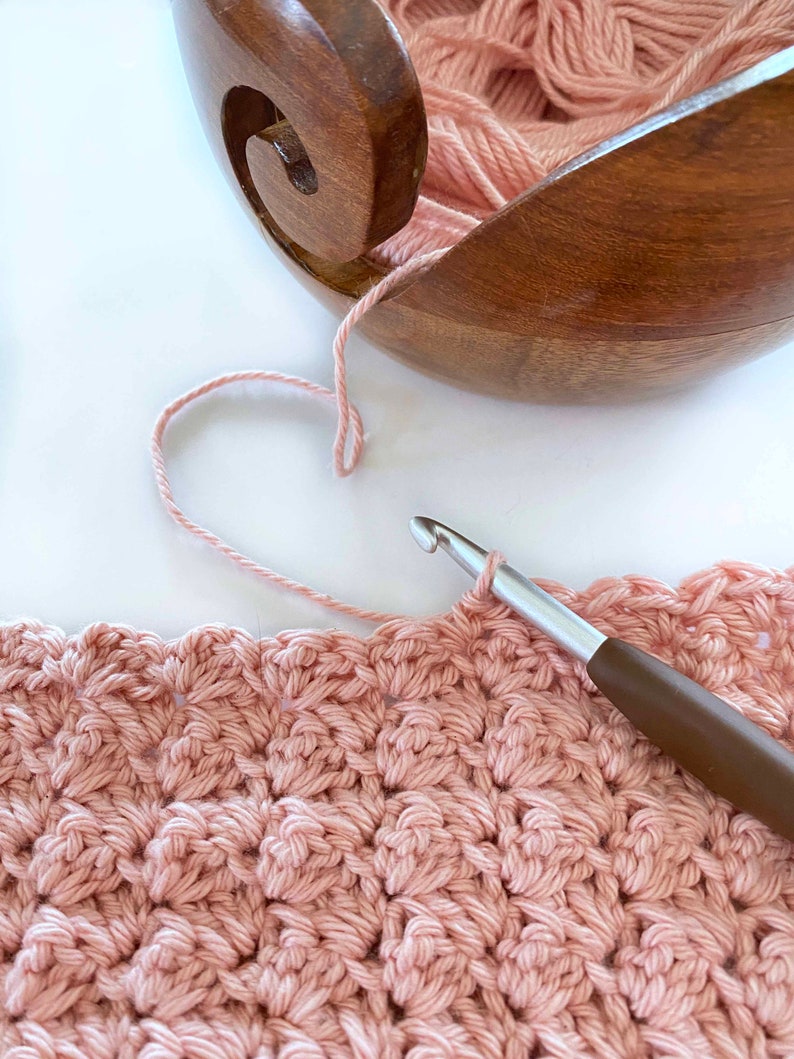 May include: A close-up of a pink crocheted blanket being worked on with a crochet hook and yarn. The yarn is coming from a wooden yarn bowl.