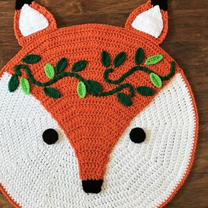 May include: A crocheted fox rug with a white background and orange face. The fox has green leaves and vines around its head and black eyes and nose.