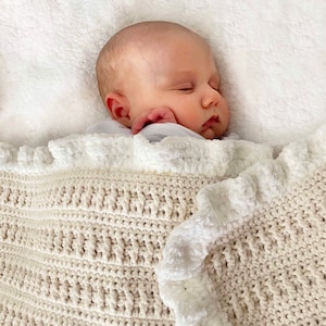 Può includere: Un bambino che dorme avvolto in una coperta all'uncinetto color crema con un bordo bianco a balze.